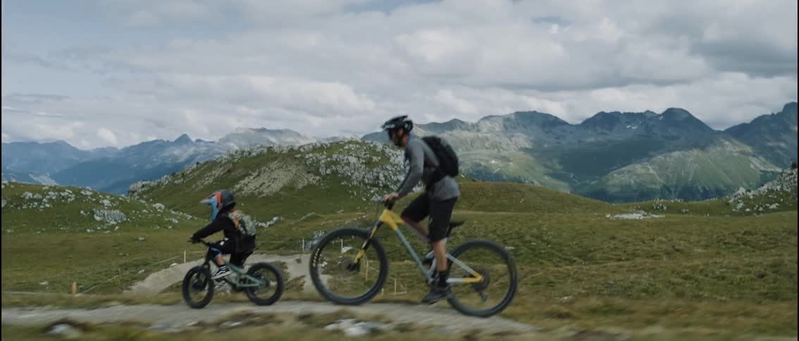 A father and his young son ride their mountain bikes in slow motion along a scenic trail through a vast alpine landscape
