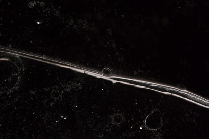 A strange, clear, viscous liquid slowly moves and morphs across a dark, textured surface