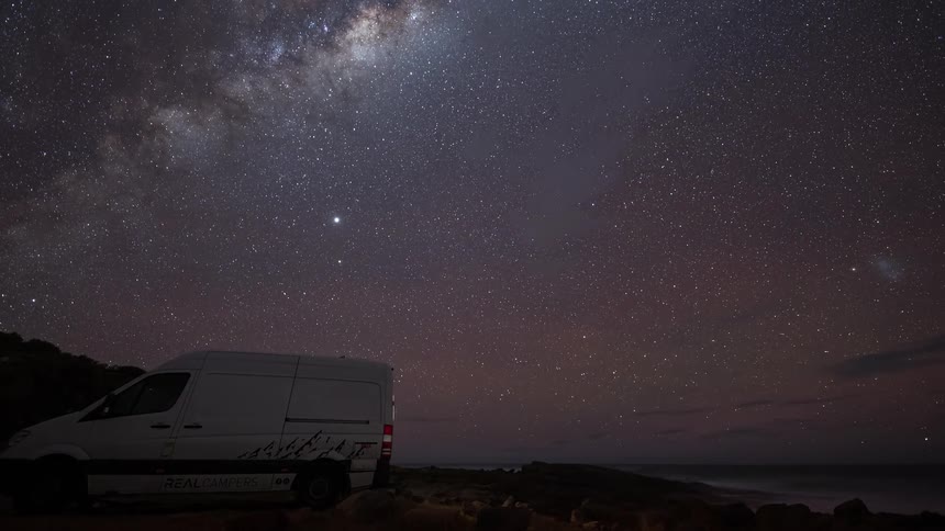 A timelapse of the Milky Way galaxy moving across the starry night sky above a camper van parked on the coast