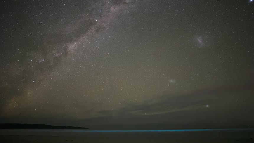 A timelapse of the Milky Way and Magellanic Clouds moving across the night sky above a beach with glowing bioluminescent waves