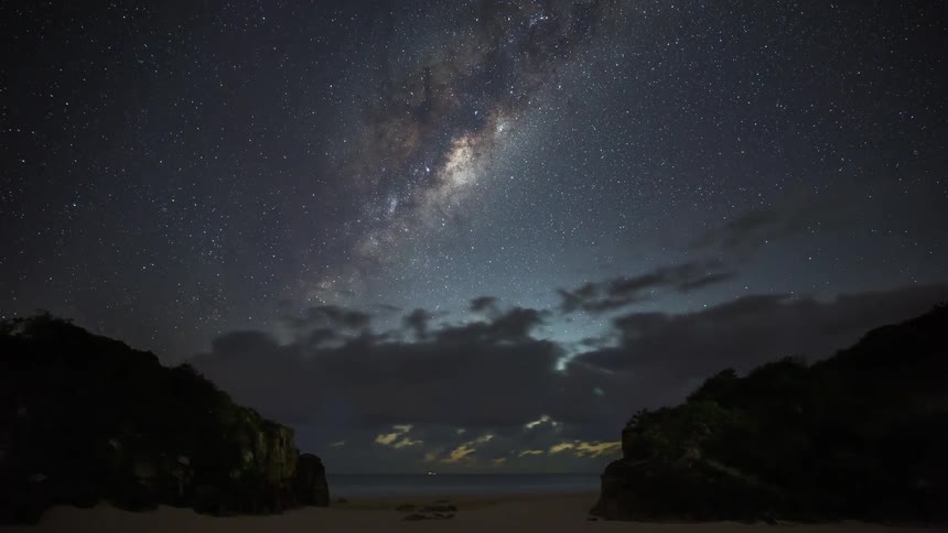 A stunning time-lapse captures the Milky Way setting over a beach as clouds roll in for a dramatic sunrise over the ocean