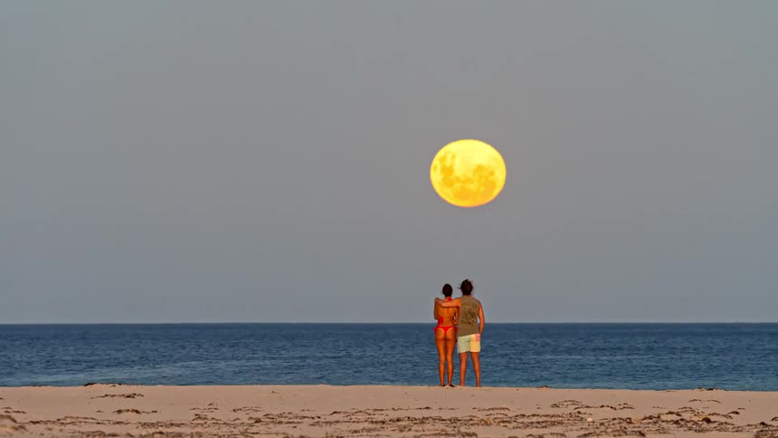 A romantic couple performs acro-yoga and watches a spectacular supermoon rise over the ocean on a sandy beach