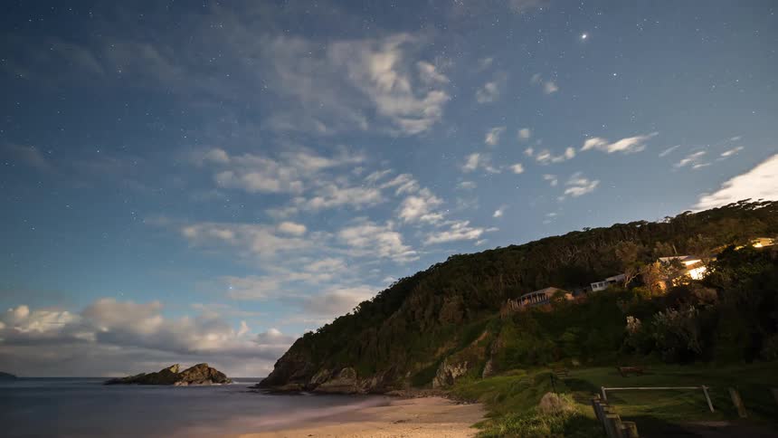 A stunning time-lapse captures the Milky Way and stars transitioning to a cloudy, moonlit night over a tranquil beach cove with illuminated homes on a hillside