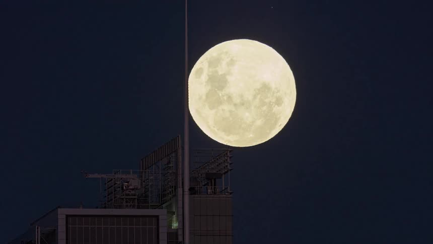 A dramatic slow tilt shot captures a large full moon ascending behind a modern skyscraper at night, eventually bisected by its spire