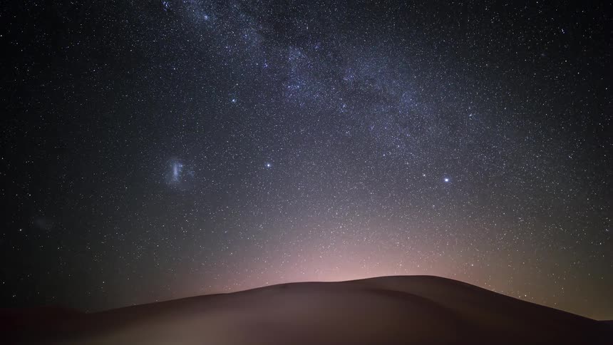 A stunning timelapse of the Milky Way and Magellanic Clouds rising in the starry night sky over desert sand dunes