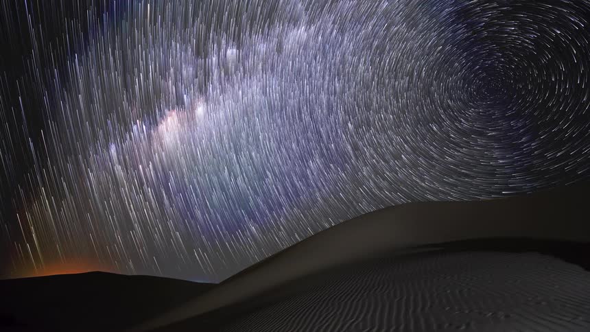 A mesmerizing time-lapse captures the Milky Way galaxy transforming into spectacular swirling star trails above tranquil desert sand dunes