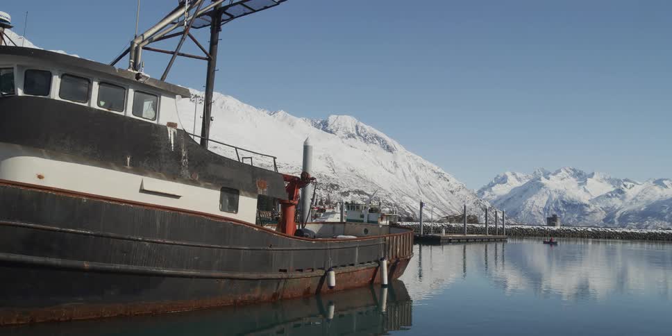 A rustic fishing boat is docked in a calm winter harbor, with a stunning backdrop of snow-capped mountains reflecting in the still water