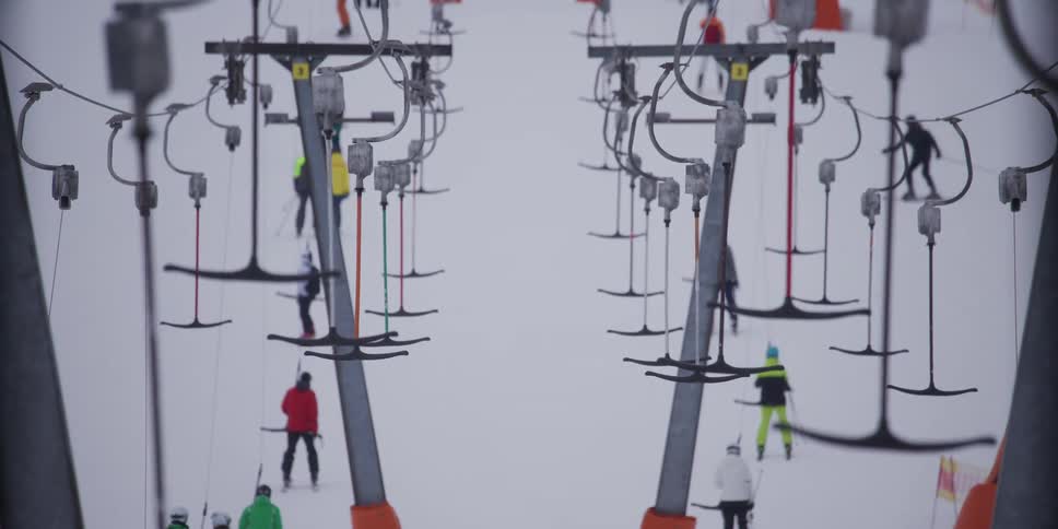 A detailed slow-motion shot shows a T-bar ski lift carrying skiers and snowboarders up a busy mountain slope