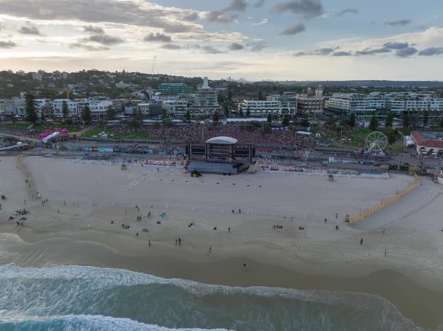 Aerial timelapse of a massive music festival with a large crowd gathered on the beach next to the ocean waves and a cityscape in the background