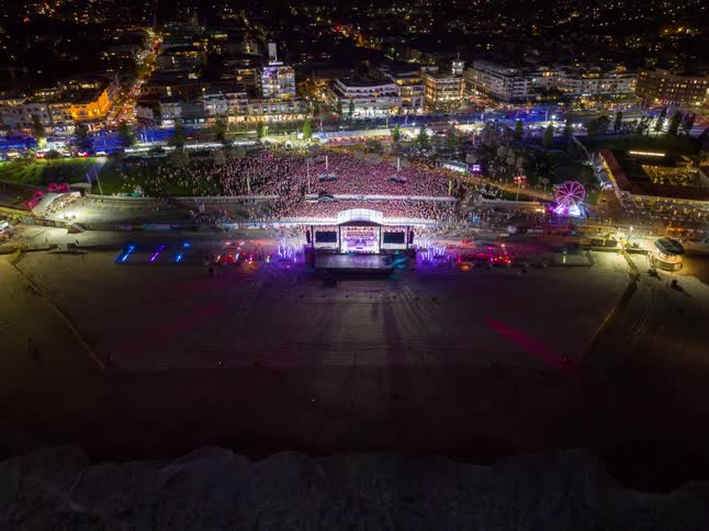 A vibrant aerial time-lapse captures a massive music festival on a beach at night, with colorful stage lights and a glowing city in the background as the camera pulls back