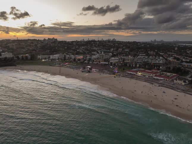 Slowly flying in from the ocean, an aerial view captures a vibrant music festival lighting up the famous Bondi Beach at sunset with the Sydney skyline in the distance