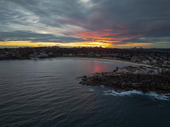 An aerial time-lapse captures a fiery orange sunset reflecting on the water as twilight descends over the iconic Bondi Beach and Sydney skyline