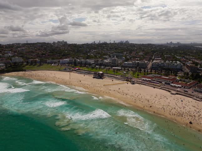 An aerial view slowly pushes in over the turquoise waves towards the crowded sands of Bondi Beach, Australia