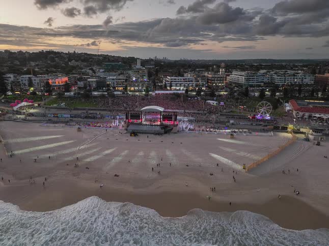 A stunning aerial time-lapse of a massive music festival on a beach at dusk, with vibrant stage lights illuminating the sand and waves crashing ashore