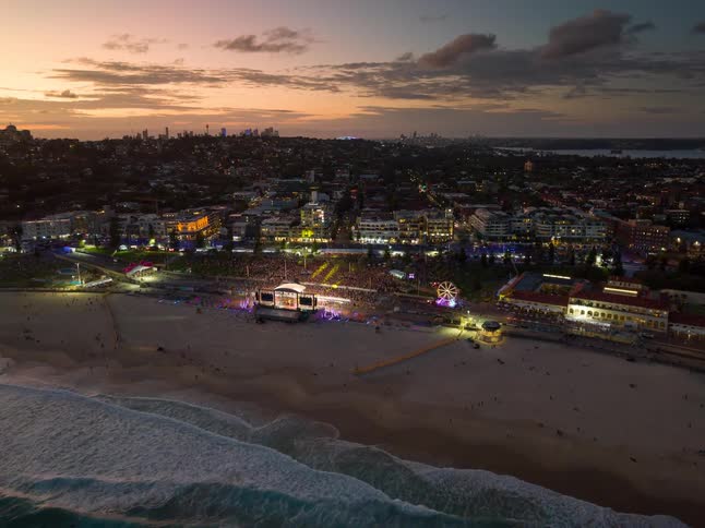 An epic aerial time-lapse captures a vibrant music festival on a crowded beach as the sun sets and the city lights begin to glow
