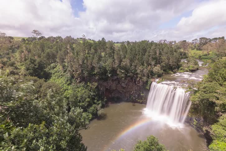 A wide, long-exposure timelapse of a majestic waterfall surrounded by a lush, windy forest, with a rainbow forming in the mist below
