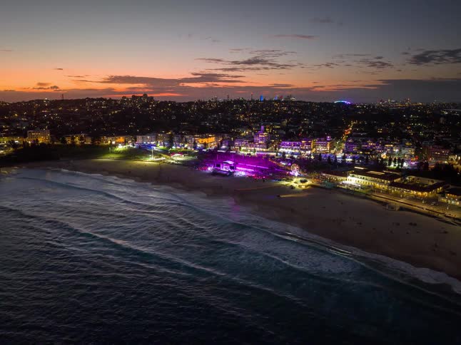 An aerial timelapse zooms in on a vibrant music festival on the beach at dusk, with colorful lights illuminating the stage as waves crash on the shore