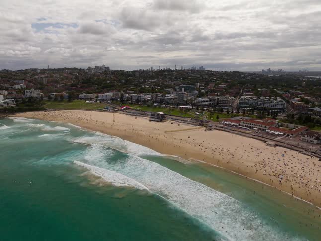 A wide aerial pullback shot reveals the famous and crowded Bondi Beach with the Sydney city skyline visible in the distance on a cloudy day