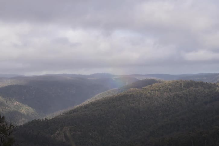 A time-lapse captures a faint rainbow fading away as shadows and clouds move over a vast forested mountain range