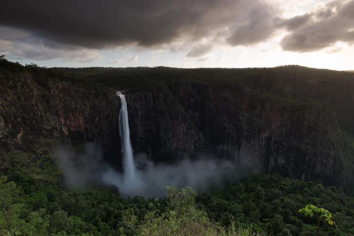 Dramatic timelapse of a powerful waterfall cascading into a misty forest canyon as storm clouds part to reveal the setting sun