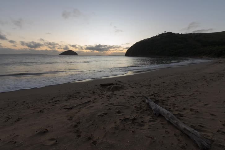 A serene timelapse captures the transition from a purple pre-dawn sky to a glorious sunrise over a secluded tropical beach and island