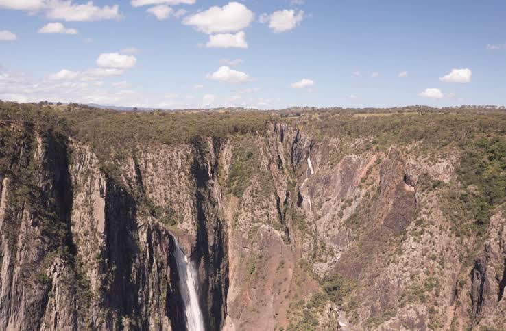 A slow aerial pullback reveals multiple waterfalls cascading into a vast, rugged canyon under a clear blue sky