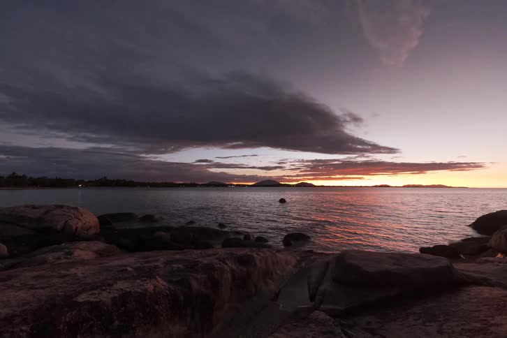 A stunning day-to-night timelapse shows a vibrant sunset fading into a starry sky over a calm, rocky bay with distant city lights
