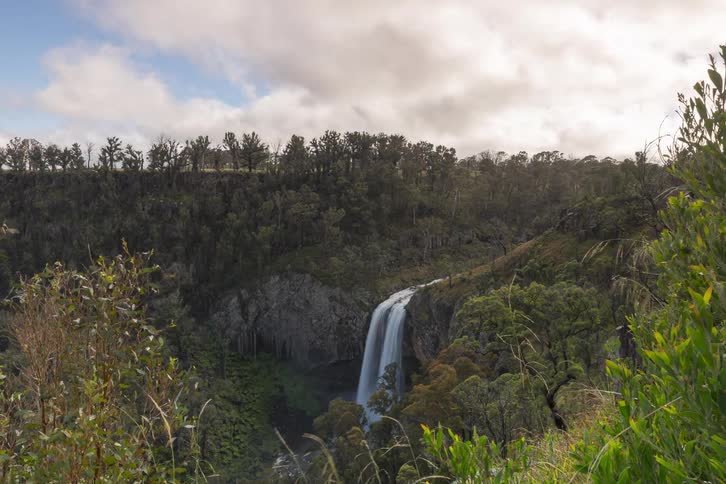 Time-lapse of clouds passing over a scenic waterfall nestled in a lush green valley, with shadows moving across the landscape