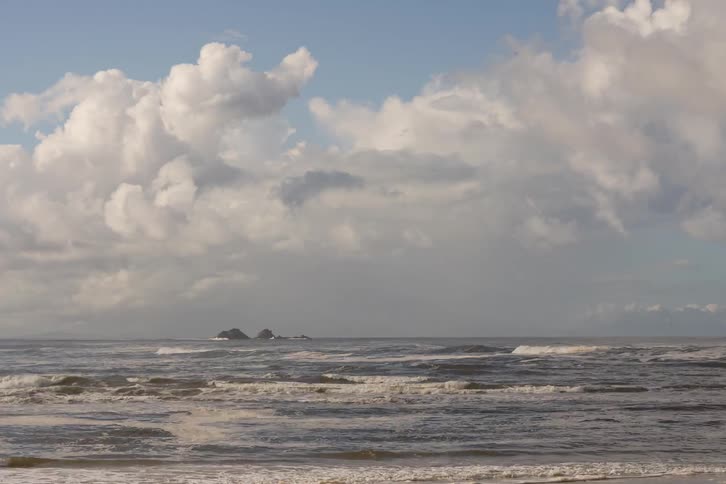 Rolling ocean waves crash on the shore under a vast, cloudy sky with distant islands on the horizon