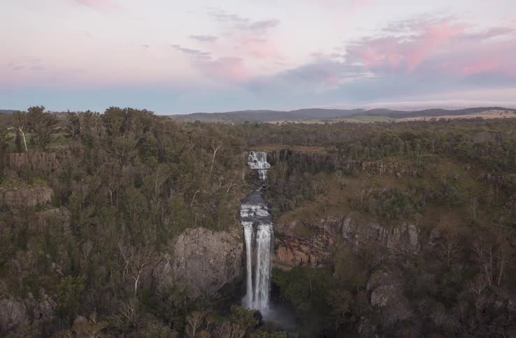 An aerial shot pulls back to reveal a stunning tiered waterfall cascading through a forested gorge at sunset