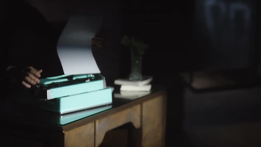 A person's hands slowly remove a finished page from a vintage typewriter in a dark, mysterious room