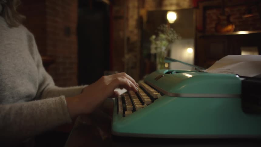 A writer's hands type on a vintage turquoise typewriter in a warmly lit, moody room