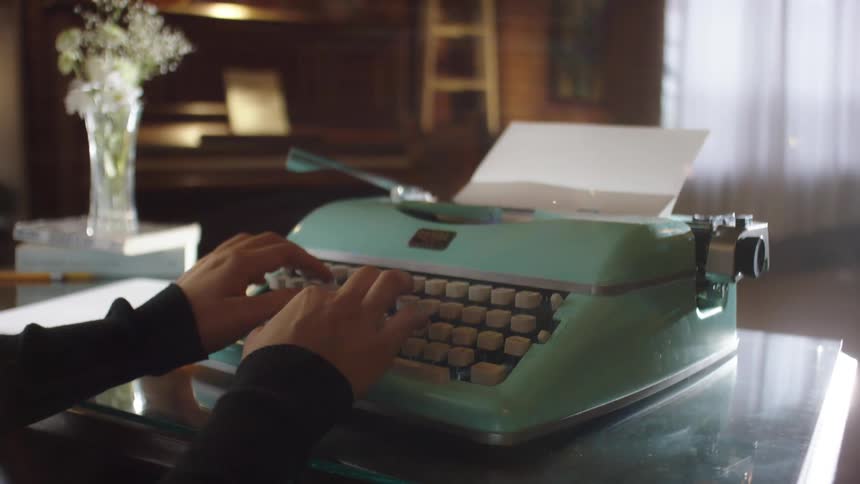 A writer types a manuscript on a vintage teal typewriter in a softly lit, cozy room