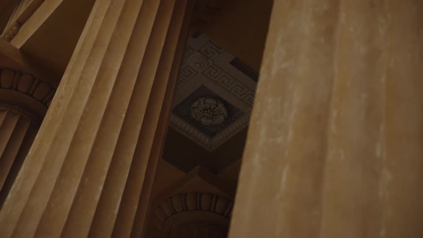 A slow zoom reveals an ornate, classical ceiling viewed between two imposing fluted pillars