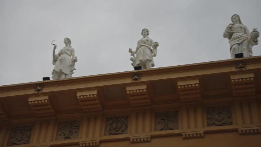 A slow pan reveals several classical white statues standing on the roof of an ornate building under a grey, overcast sky