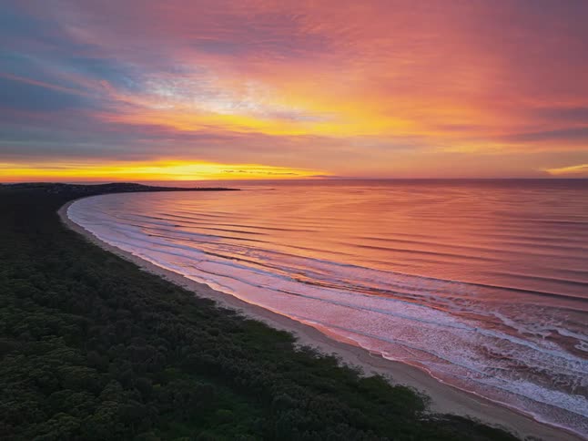 An incredible time-lapse captures a fiery red and orange sunrise reflecting on the ocean as waves roll onto a forested coastline