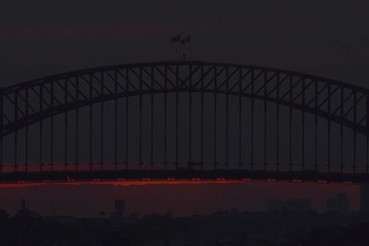 A striking timelapse captures the large red sun setting behind the iconic Sydney Harbour Bridge as a train crosses