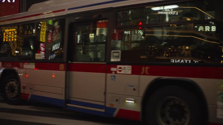 A city bus slowly drives through a crosswalk at the bustling Shibuya Crossing in Tokyo at night, with crowds waiting on the sidewalk