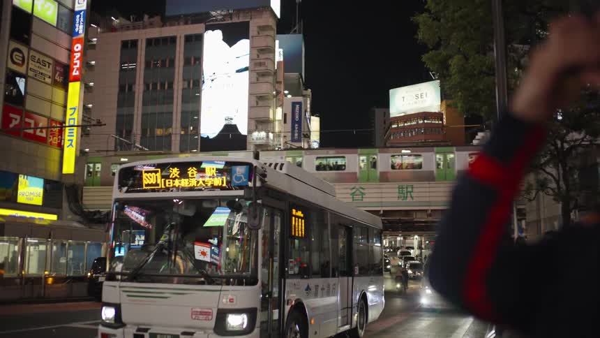A city bus waits at a busy intersection as a train passes on an elevated track in the brightly lit Shibuya district of Tokyo at night