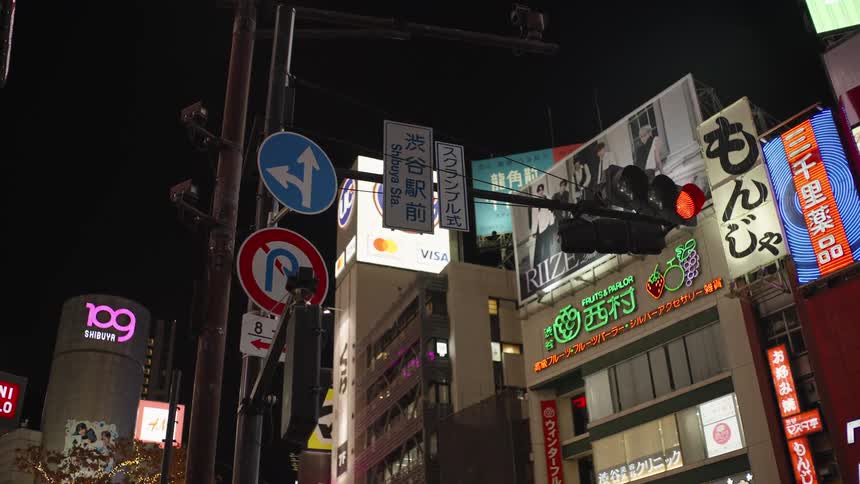 A vibrant street corner in Shibuya, Tokyo, pulses with energy at night, illuminated by glowing neon signs, billboards, and a red traffic light