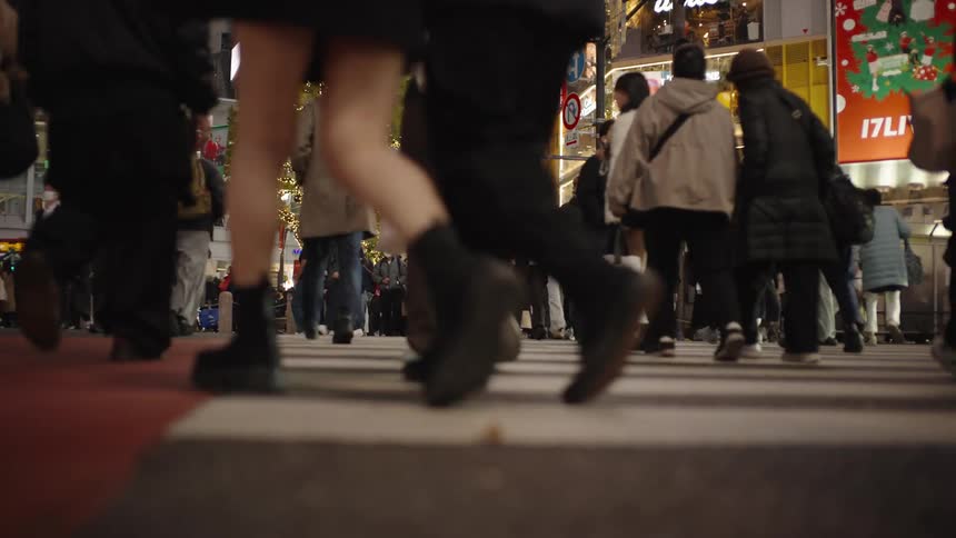 A low-angle, slow-motion shot captures the feet of pedestrians crossing a busy street in Tokyo at night