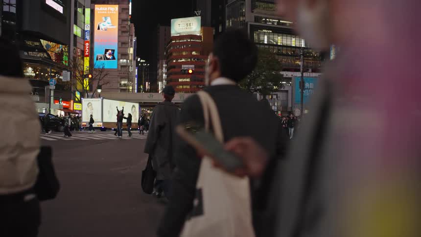 A slow-motion, over-the-shoulder view of pedestrians crossing a bustling Tokyo intersection at night, surrounded by bright city lights
