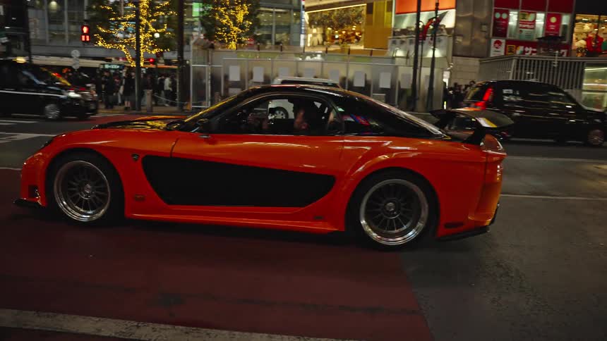 A highly modified orange and black sports car cruises through the brightly lit streets of Tokyo, Japan at night