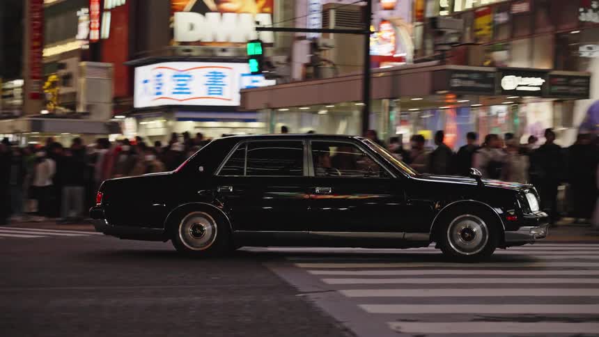 A black luxury sedan slowly cruises through a bustling crosswalk in the neon-lit city of Tokyo at night