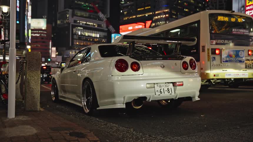 A modified white Nissan GT-R R34 sports car is parked on a bustling Tokyo street at night, with a city bus and bright lights in the background