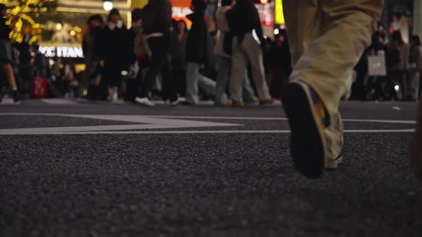 A low-angle, slow-motion view of a diverse crowd's legs and feet crossing a busy city street at night in Tokyo