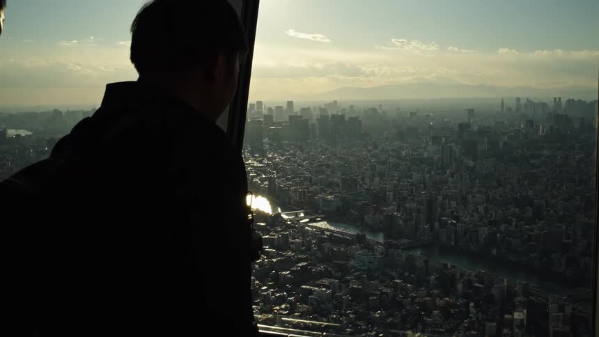 A silhouetted man with a backpack looks out a window from a skyscraper over a sprawling city at sunset