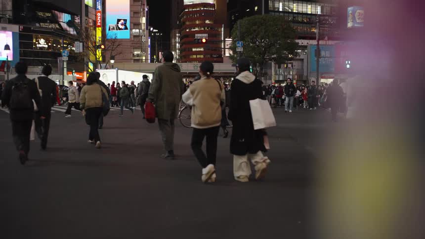 A diverse crowd of people crosses a busy city street in slow motion at night, with neon lights glowing in the background