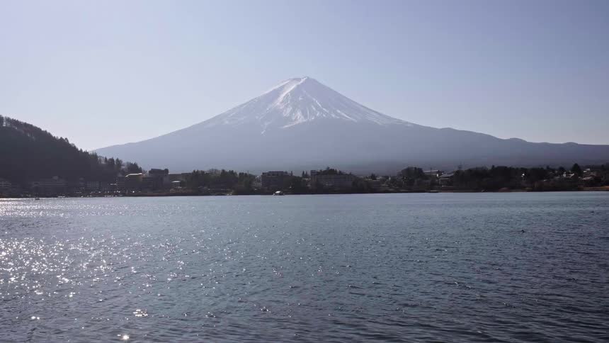 A serene and static view of the majestic, snow-capped Mount Fuji across a shimmering lake on a clear, sunny day in Japan