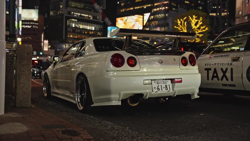 A modified white Nissan Skyline GT-R R34 is parked on the side of a busy street in Tokyo at night